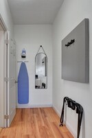 Un pasillo con suelo de madera, una puerta, una tabla de planchar azul, un espejo fijado a la pared y un estante de metal negro.