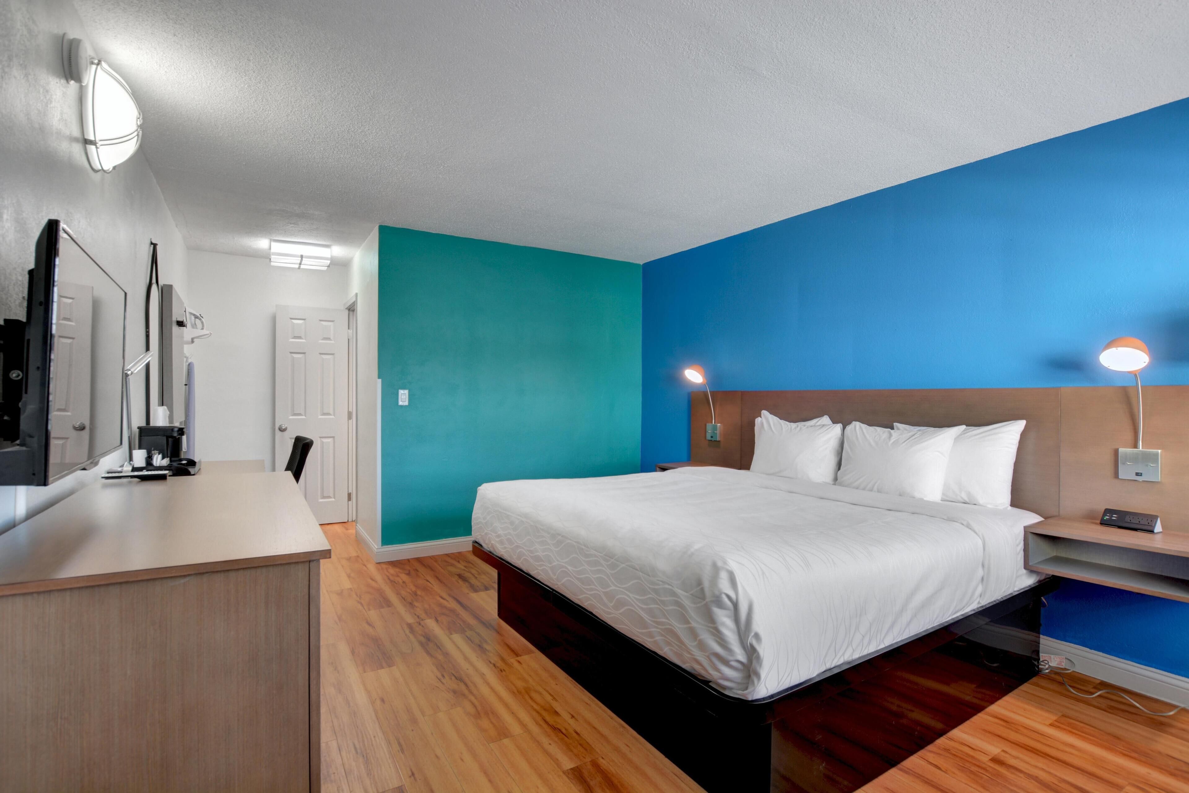 A modern hotel room with a bed, bedside table, lamp, and a desk with a chair.