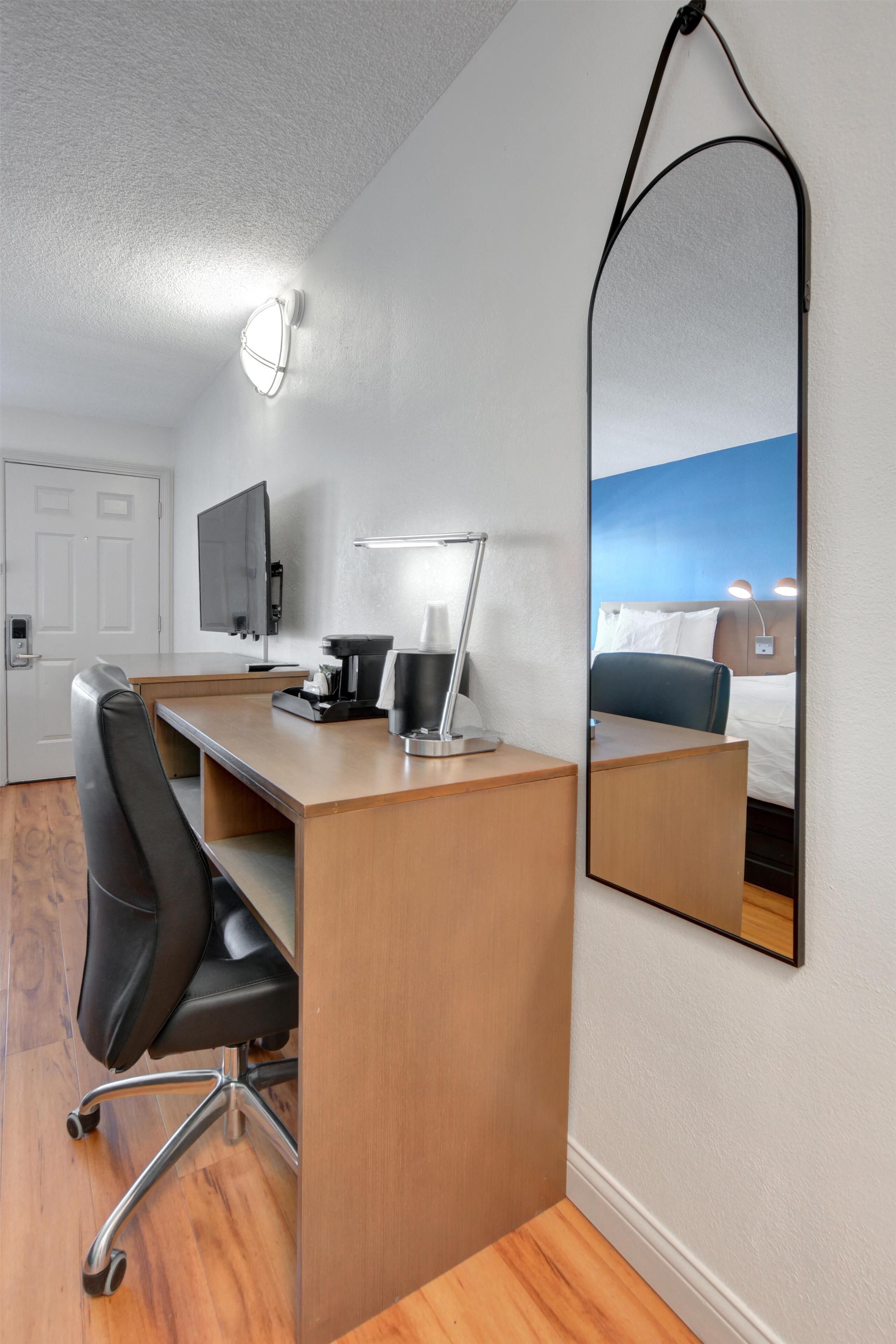 A hotel room with a desk, chair, flat-screen TV, bedside table, lamp, and a large mirror reflecting the room.