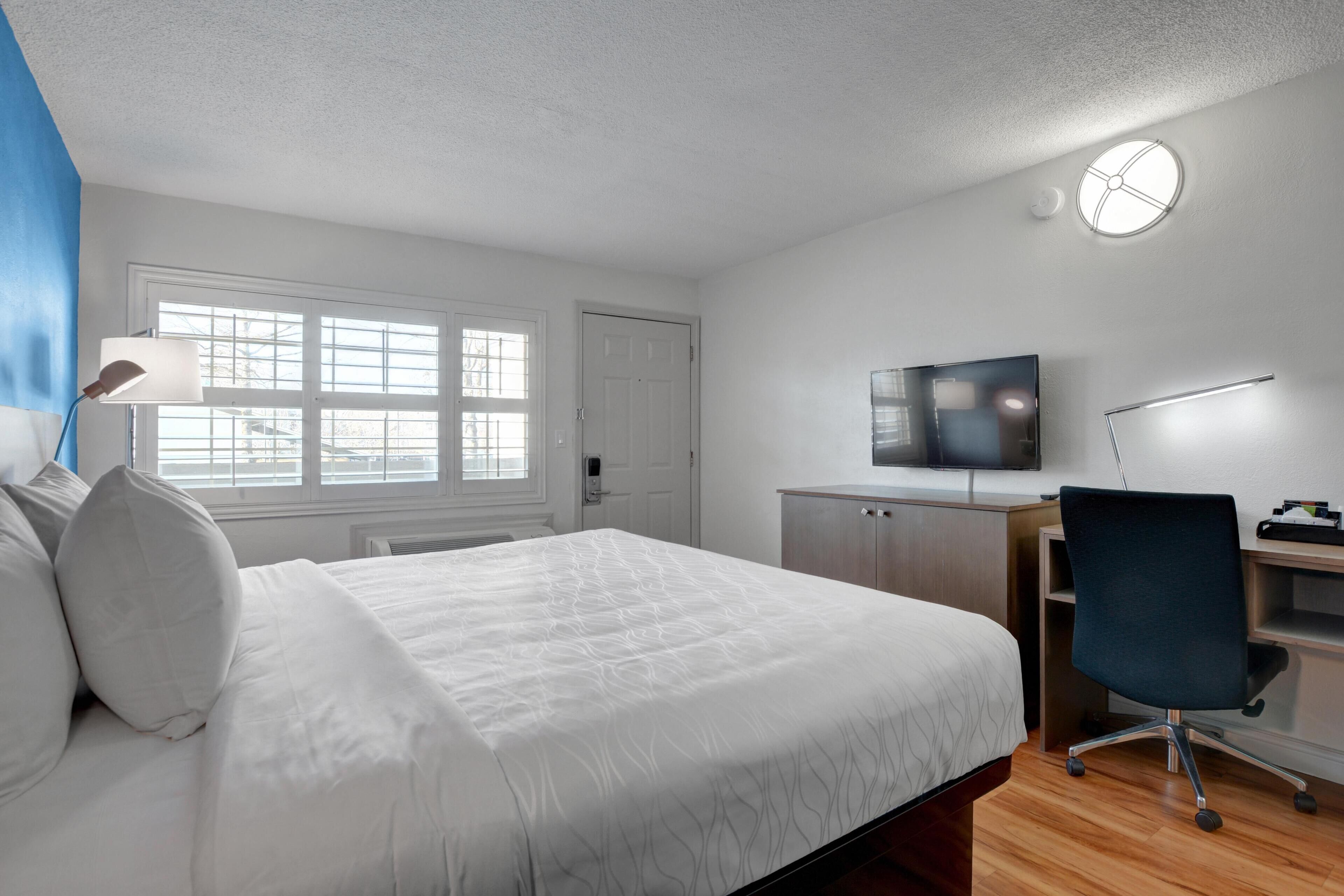 A bedroom with a bed, a desk, a chair, a television, and a window.