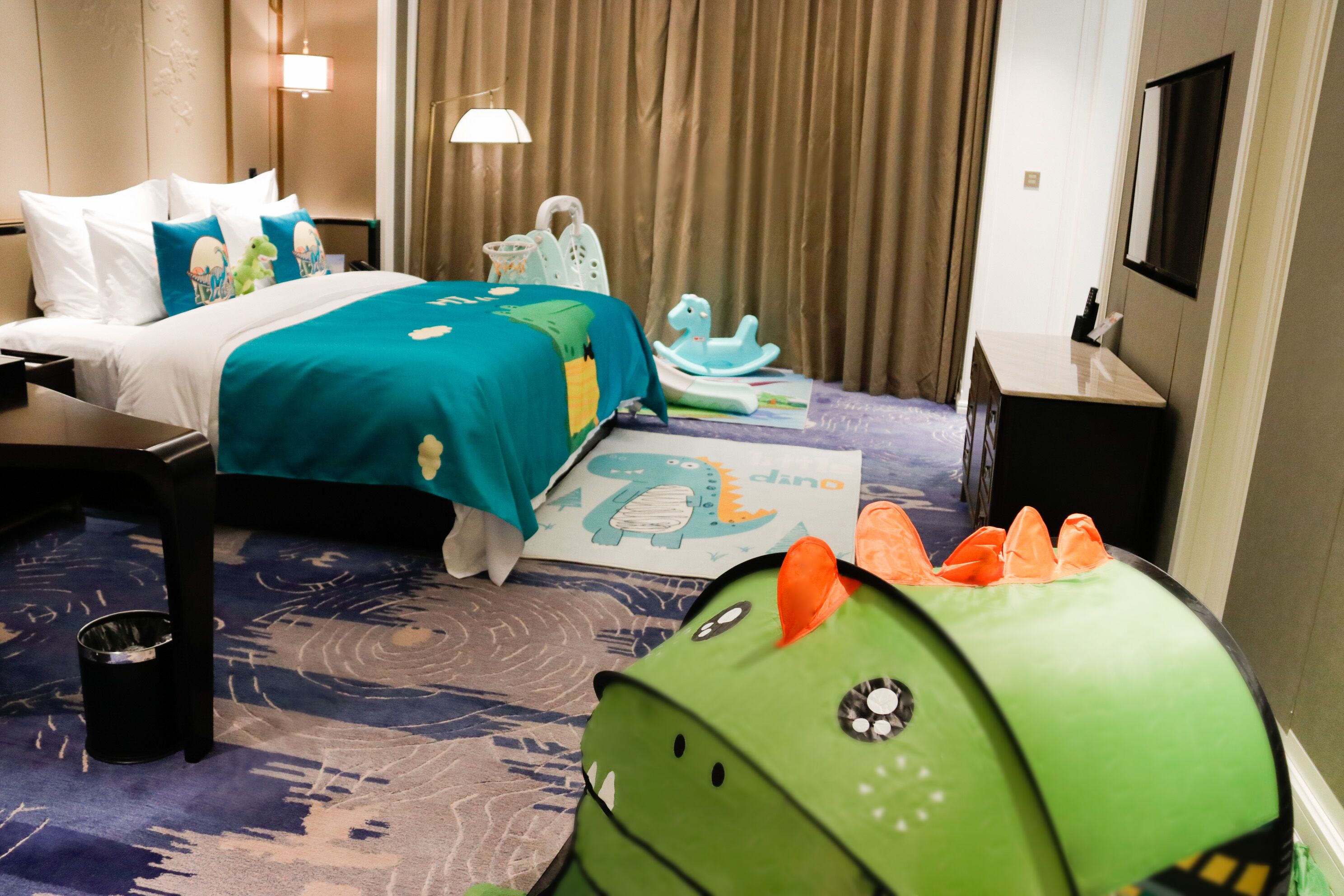 Una habitación de hotel con una cama, un escritorio, una silla, una mesita, un cubo de basura y un juguete de dinosaurio verde.