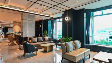 A modern hotel lobby with a spacious seating area, a coffee table, and a large window offering a city view.