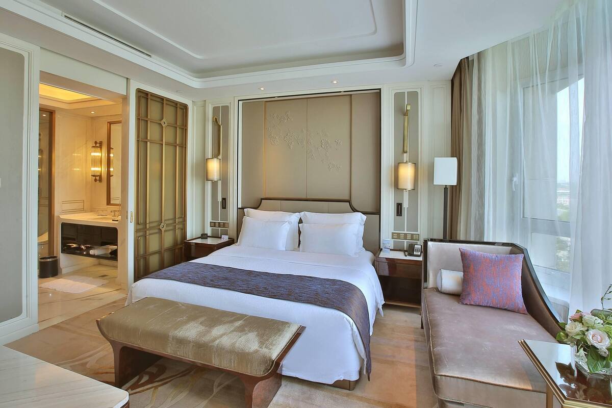 Habitación de hotel con una cama grande, un sofá, un banco y una mesita. Se ve un baño al fondo.