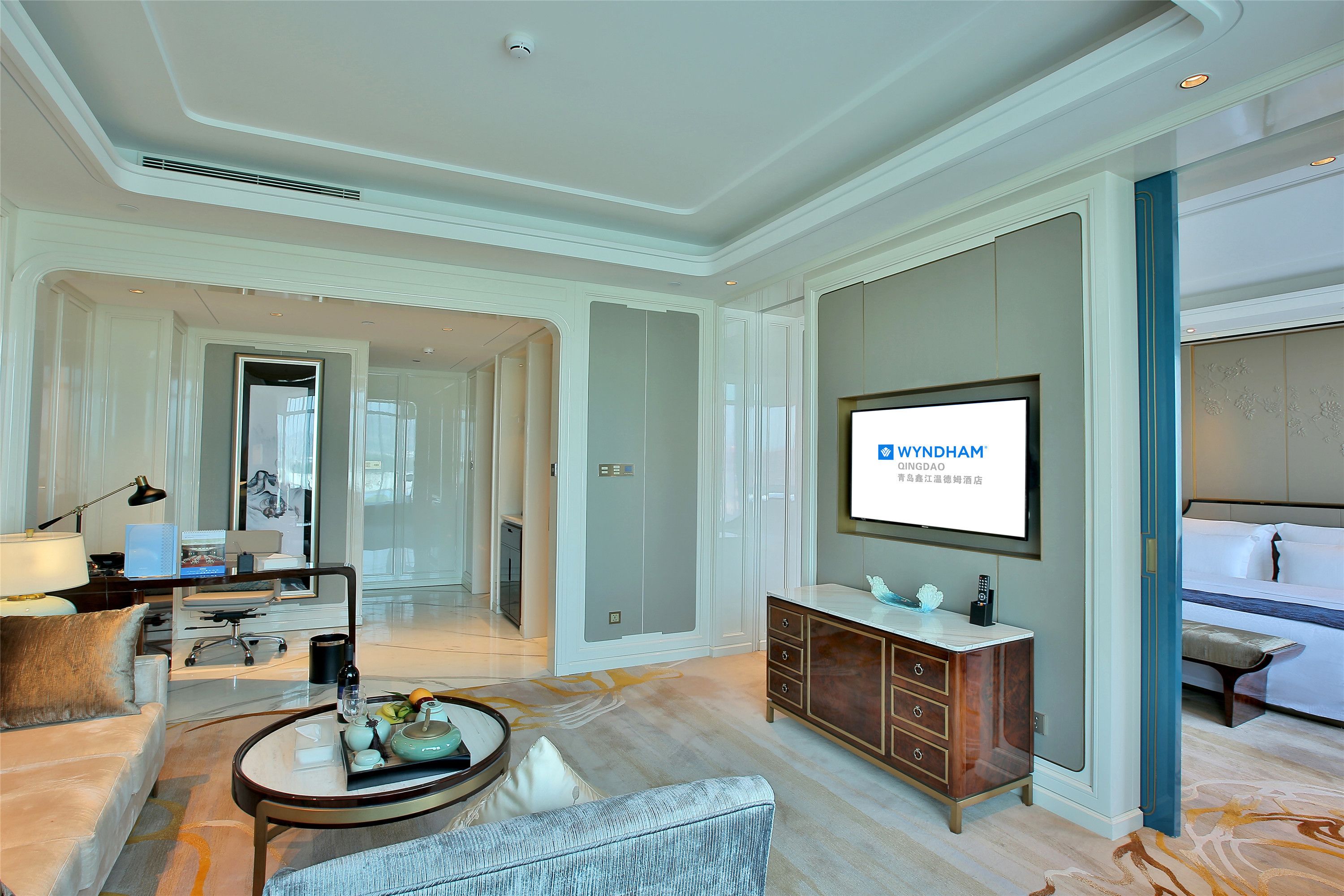 A modern hotel room with a large flat-screen TV, a comfortable seating area with a sofa and armchair, a wooden cabinet, and a large mirror.