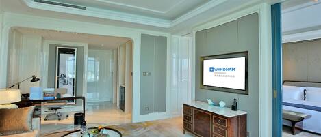 A modern hotel room with a large flat-screen TV, a comfortable seating area with a sofa and armchair, a wooden cabinet, and a large mirror.