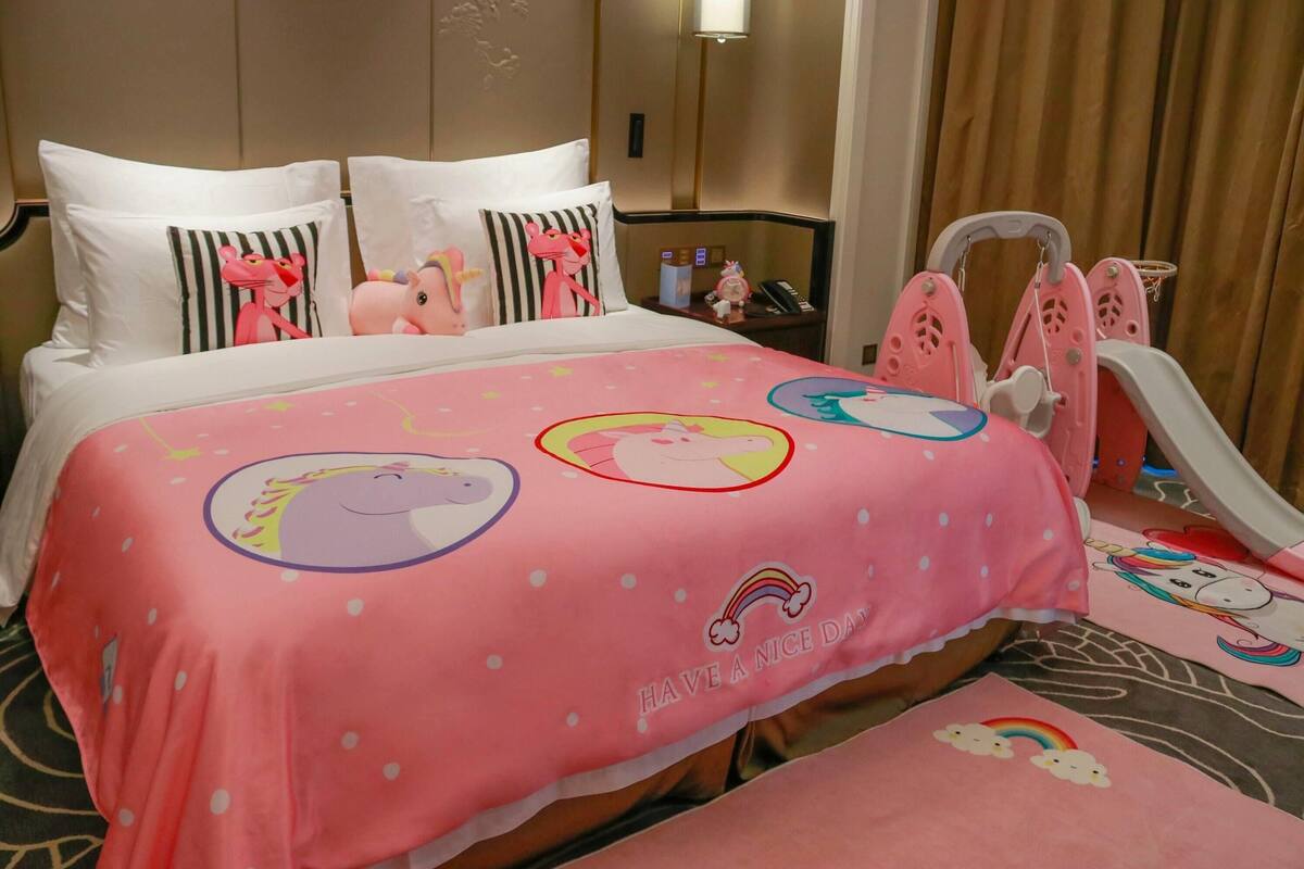 Habitación de hotel con edredón rosa con diseños de unicornios y arcoíris, una mesita de noche con lámpara y teléfono, y una alberca para juegos rosa con diseño de unicornio.