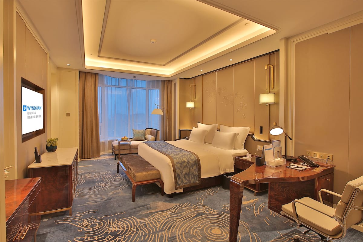 Habitación de hotel con una cama grande, un escritorio con lámpara, una silla, un banco y una pantalla de televisión mostrando 'WYNDHAM QINGDAO'.