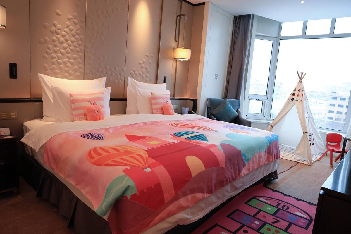 Habitación de hotel con una cama grande, un escritorio, una silla, un ventanal con vista a la ciudad y una tienda tipi.
