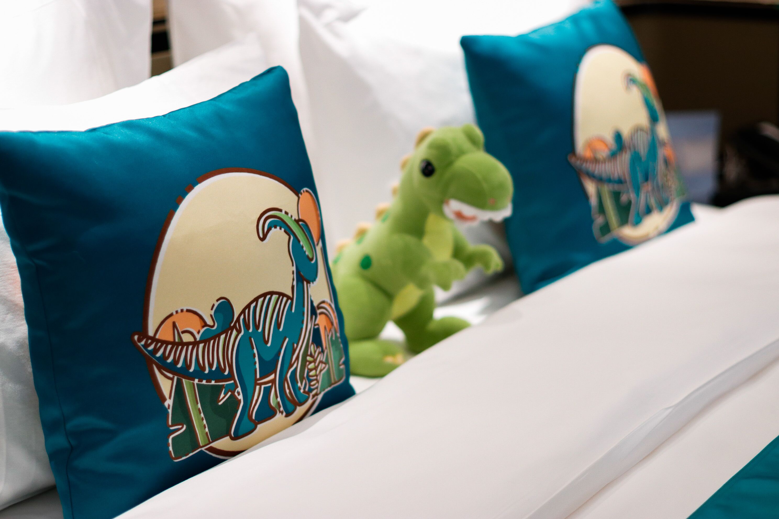 Un lit avec des oreillers de couleur turquoise ornés d’un motif de dinosaure, un jouet en peluche vert et du linge de lit blanc.