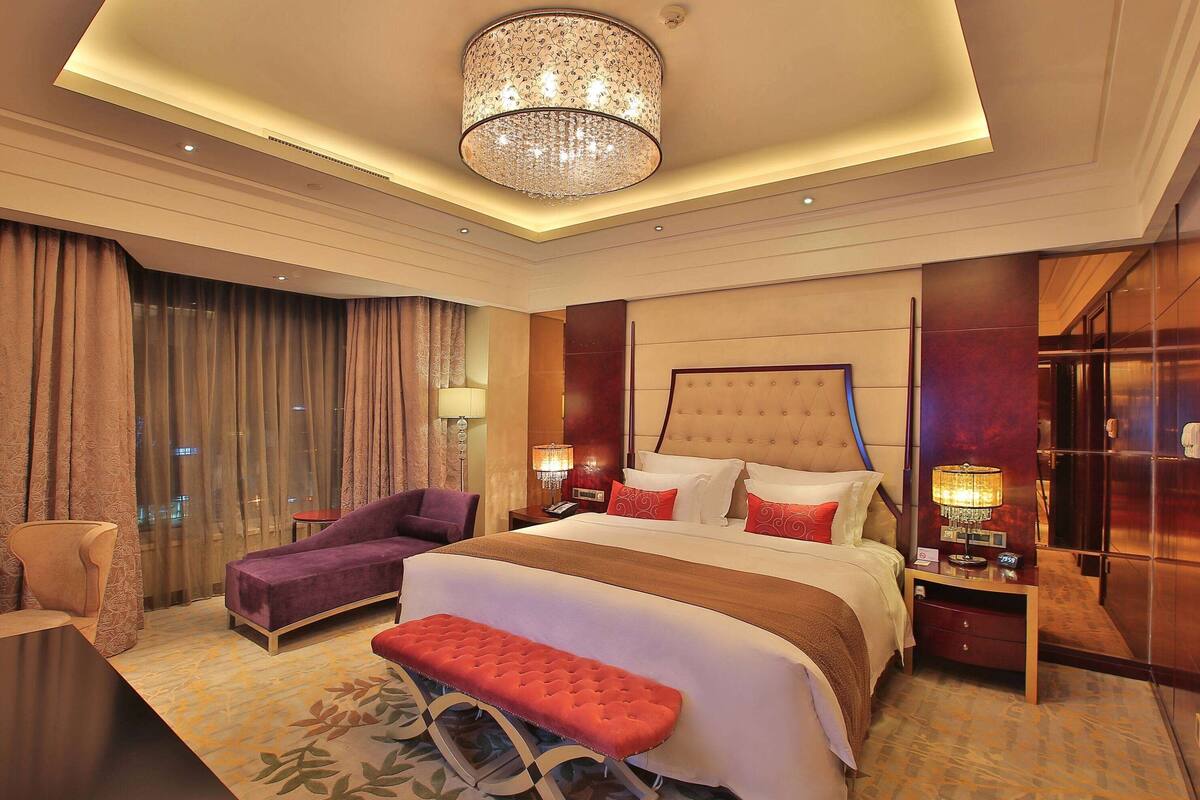 Una habitación de hotel moderna con una cama grande, un sillón, una banca, una mesita de noche con lámpara y un gran candelabro.