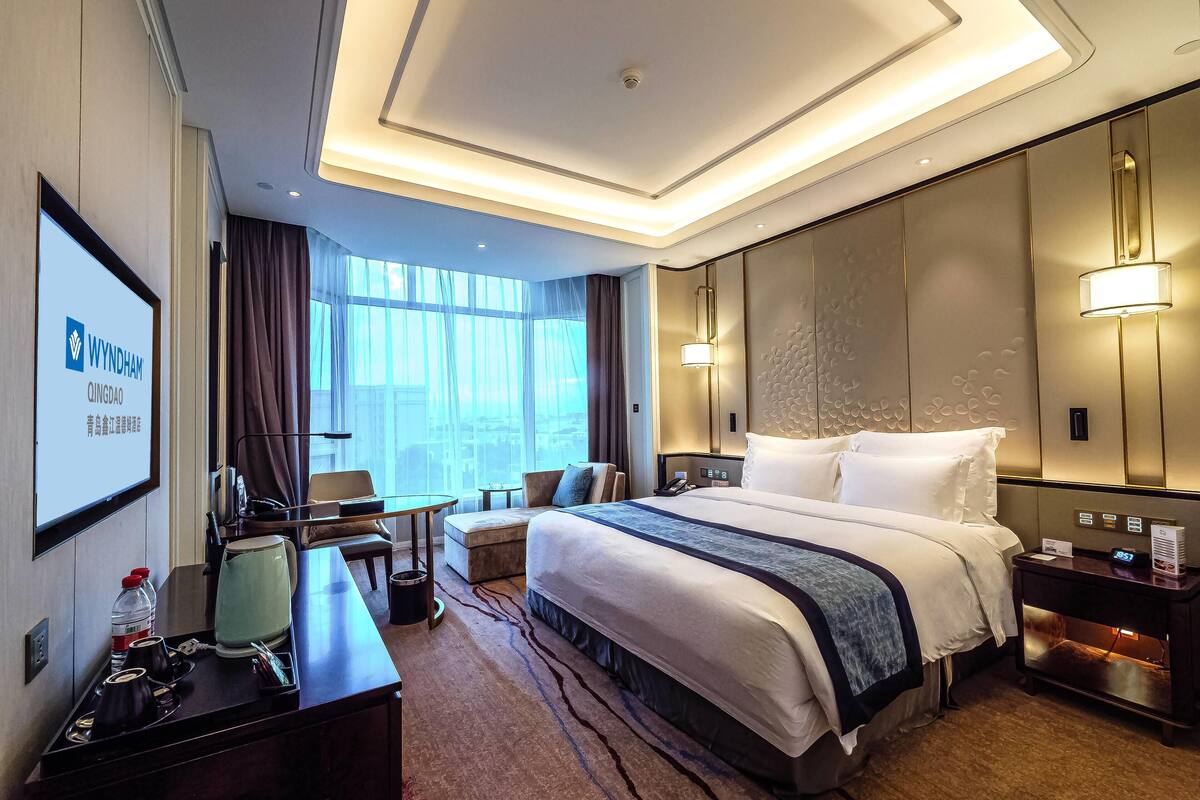 Habitación de hotel con una cama grande, un escritorio con silla, una mesita, un sofá y un ventanal con cortinas.