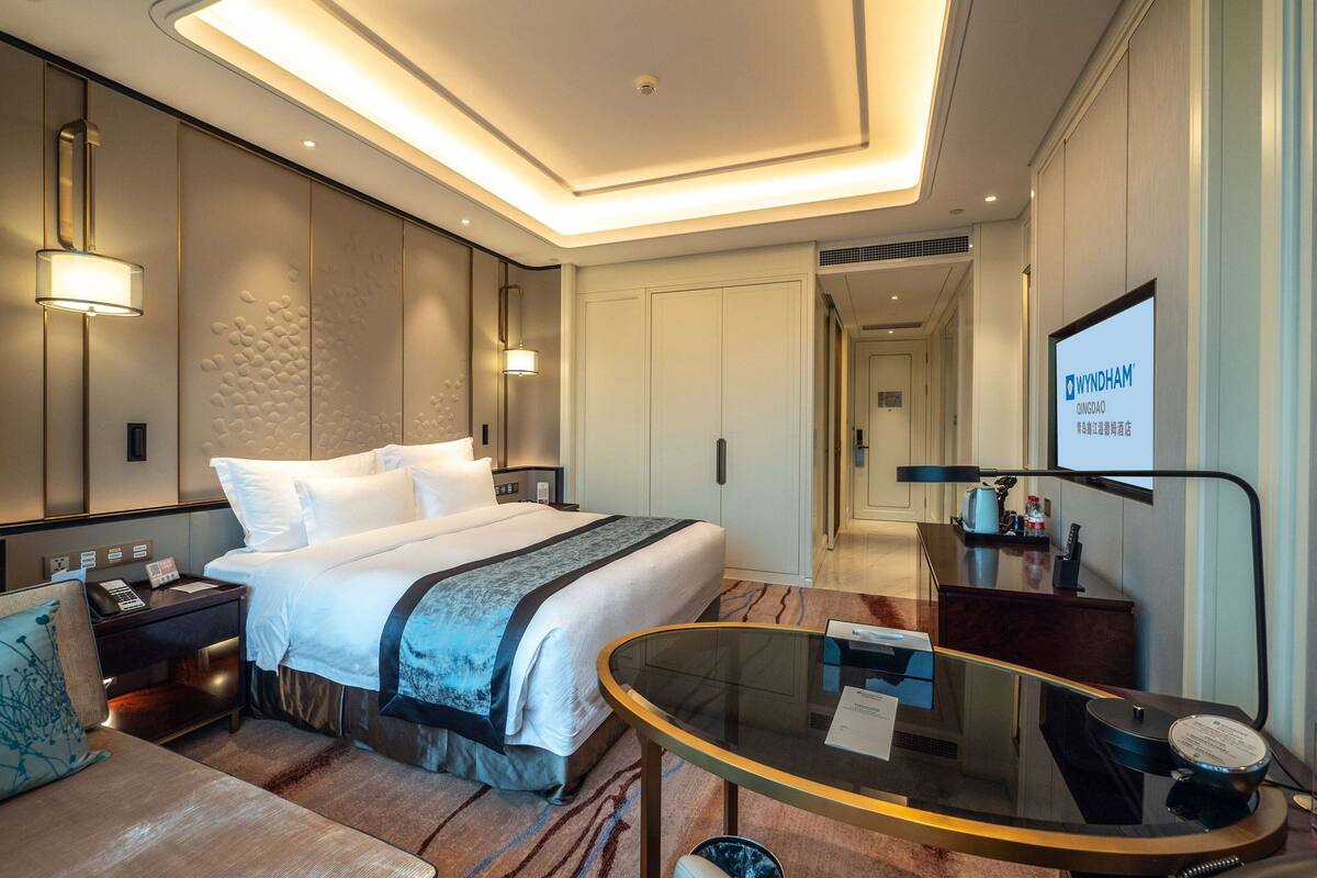 Habitación de hotel con una cama grande, mesitas de noche, un escritorio, una mesa de vidrio y un televisor mostrando 'WYNDHAM'.
