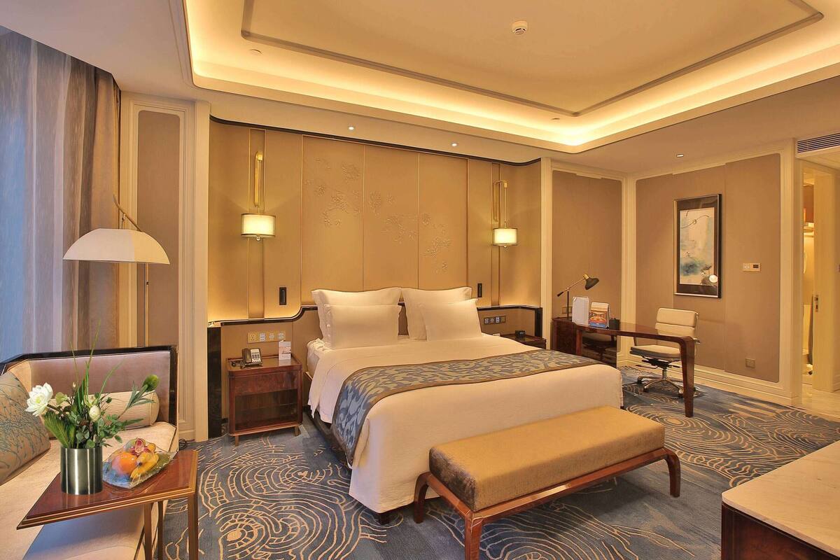Una habitación de hotel con una cama grande, mesitas de noche, un escritorio, una silla, un banco y un jarrón decorativo con flores.