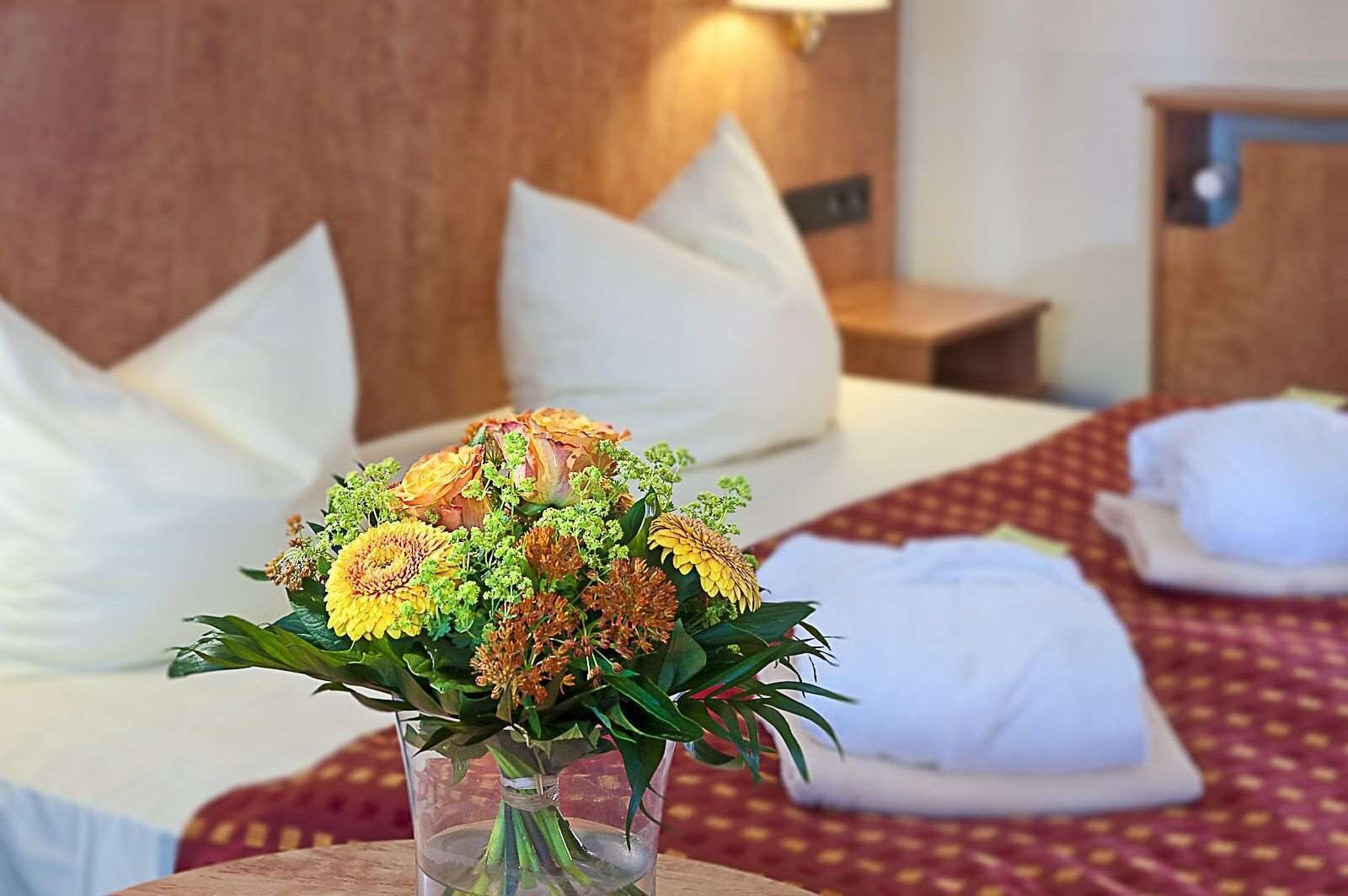Una habitación de hotel con una cama, almohadas, una mesita de noche y un jarrón con flores.