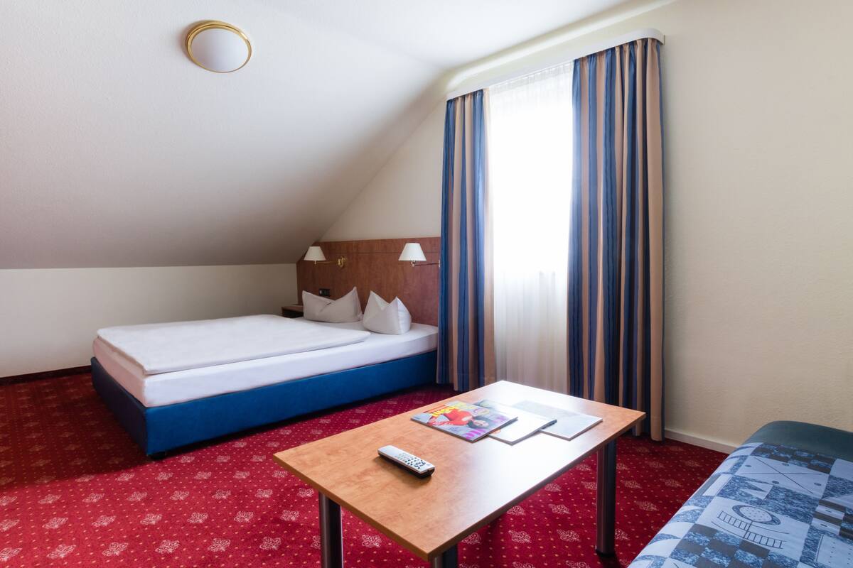 A hotel room with a single bed, a small table with a remote control, and a window with curtains.