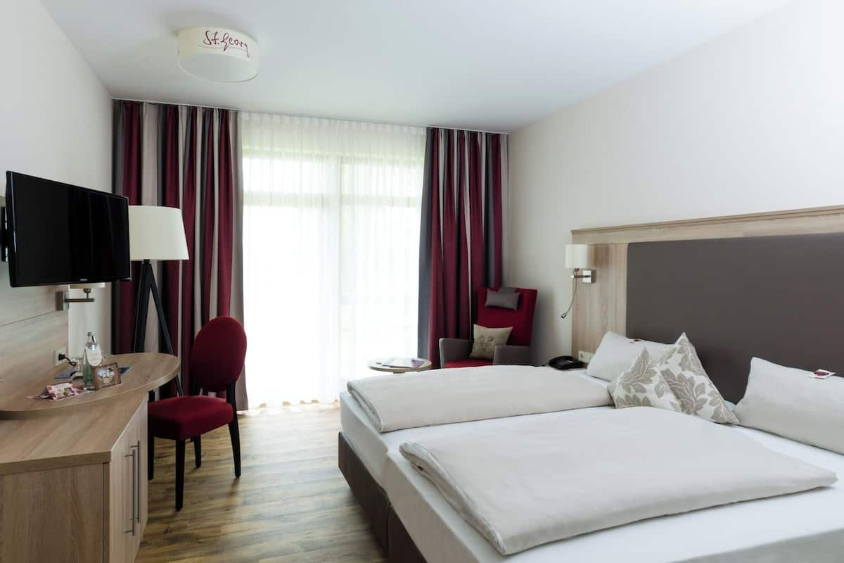 A hotel room with two beds, a desk, a chair, a TV, and a large window with curtains.