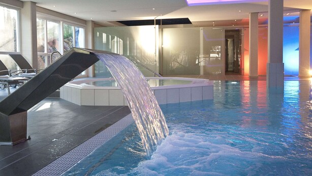 An indoor swimming pool with a waterfall feature, seating areas, and a modern architectural design.