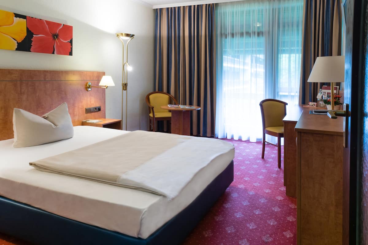 A hotel room with a large bed, a desk, a chair, a lamp, and a window with curtains.