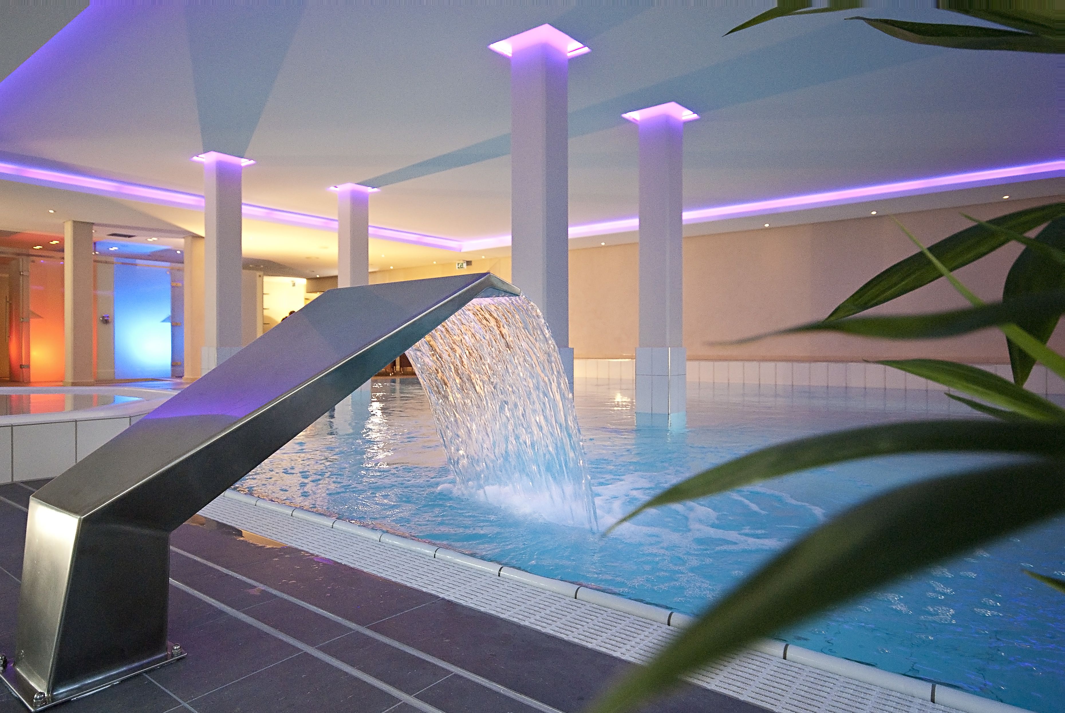 A modern indoor pool with a water feature, surrounded by white pillars and ambient lighting.