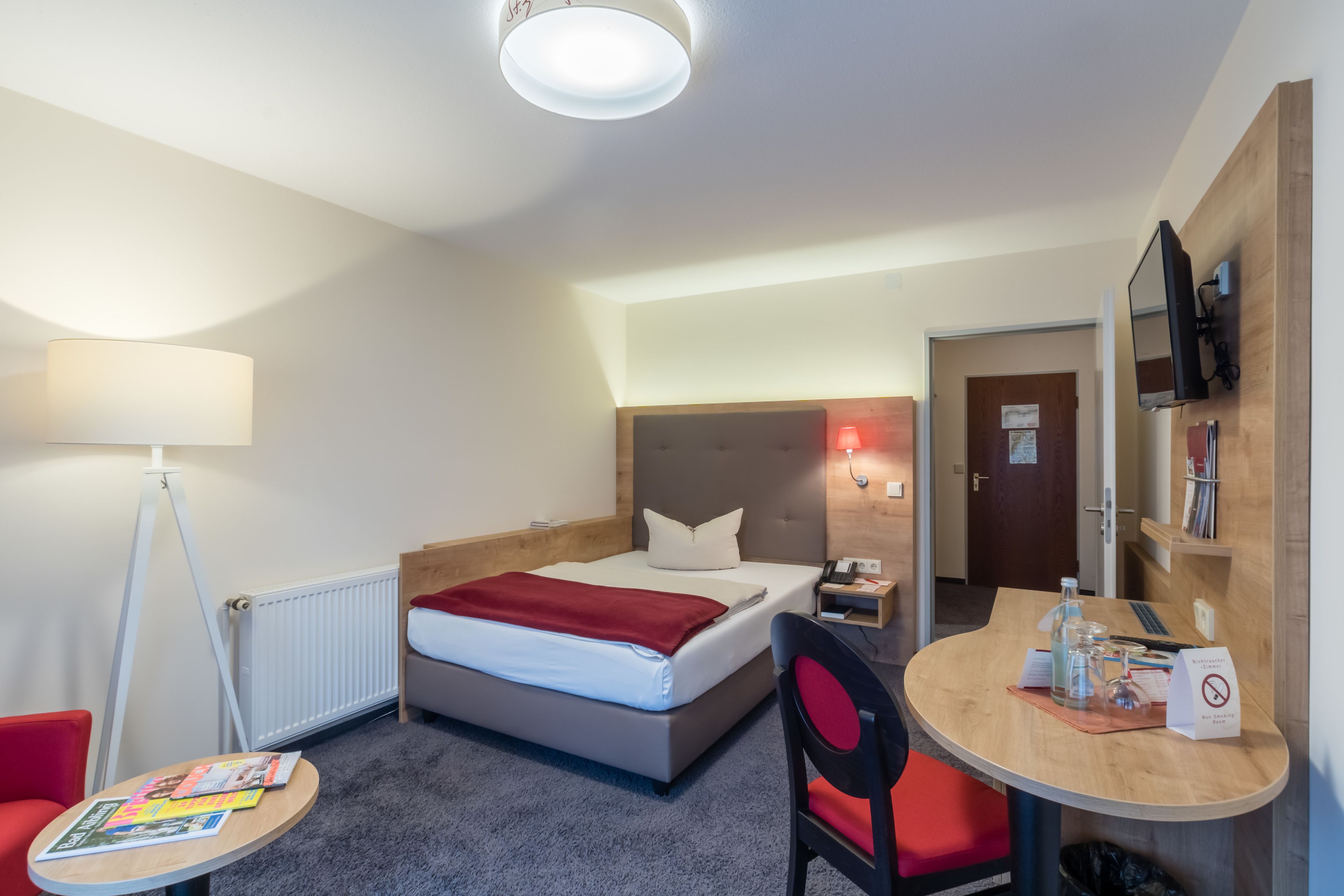 Una habitación de hotel con cama, mesita de noche, lámpara, silla y una pequeña mesa con una revista.