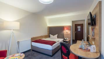 Una habitación de hotel con cama, mesita de noche, lámpara, silla y una pequeña mesa con una revista.