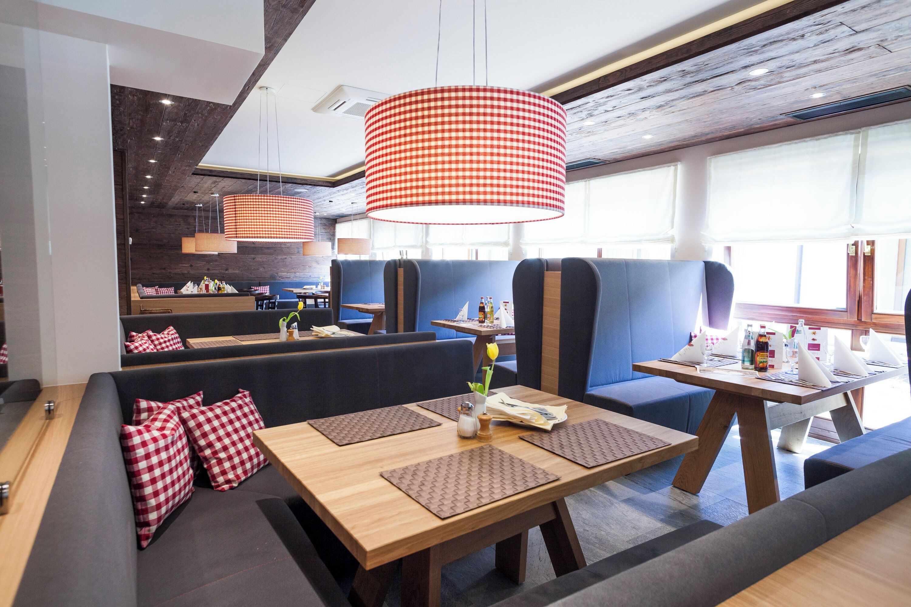 Ein modernes Restaurant mit Holztischen und blauen Sitzbänken, mit karierten Kissen und einer großen, rot-weiß karierten Pendelleuchte.