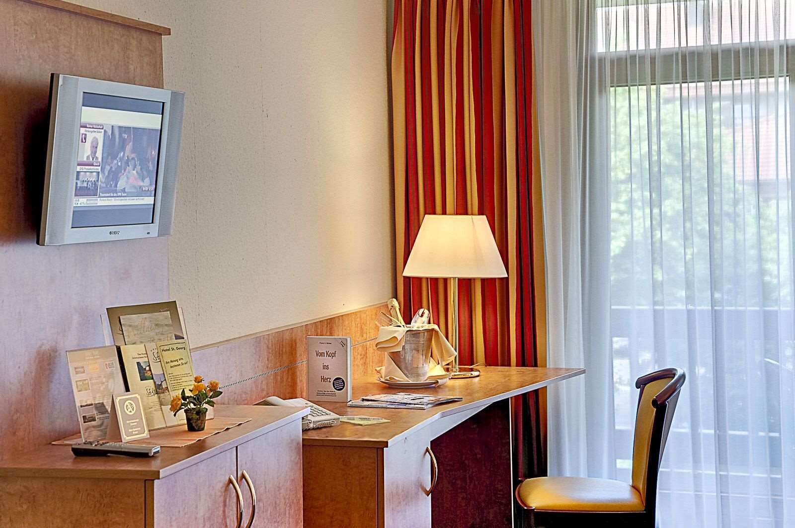 A hotel room with a desk, a television mounted on the wall, a lamp, a chair, and a window with curtains.