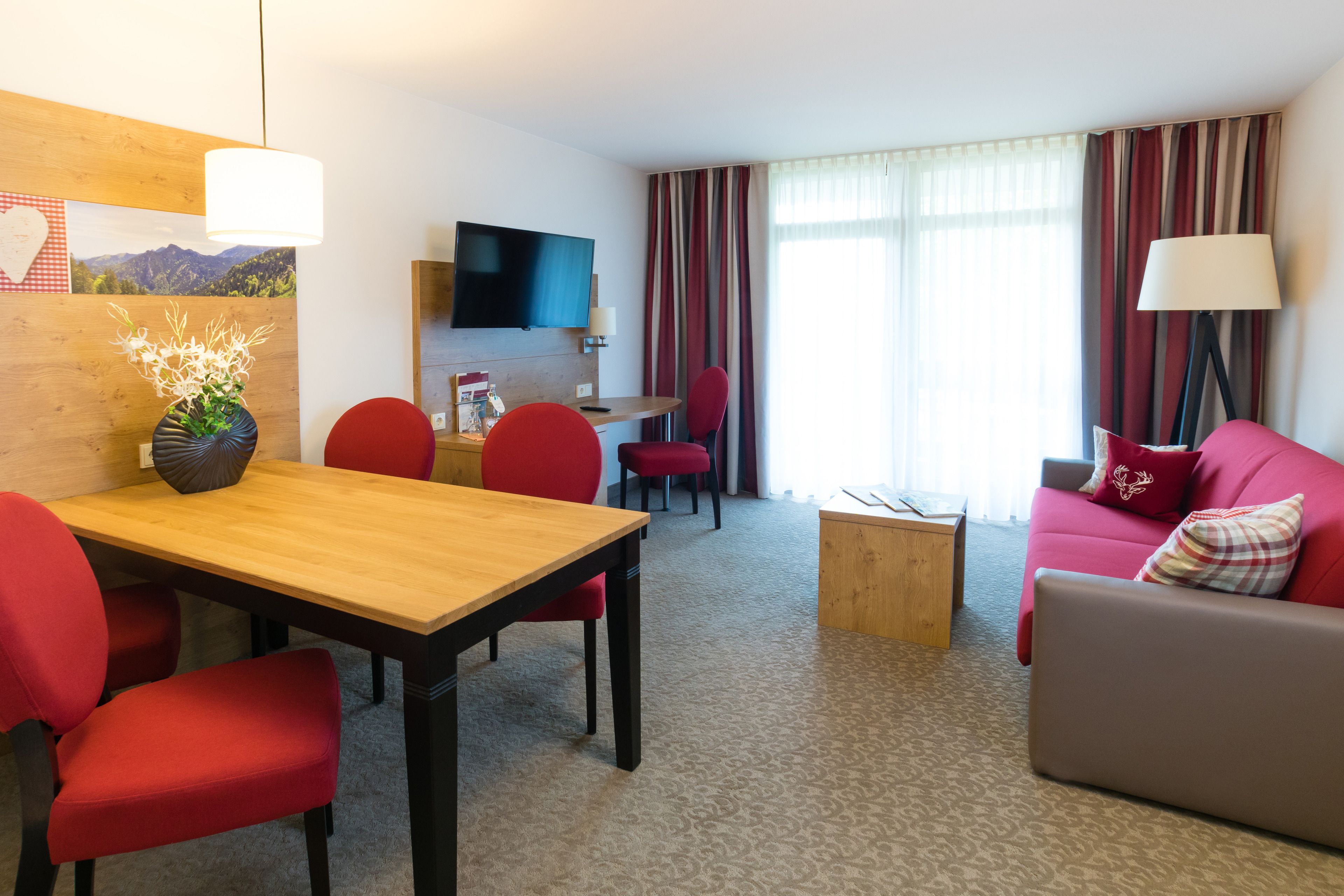 A hotel room with a wooden dining table, red chairs, a red sofa, a grey sofa, a small wooden side table, a TV mounted on the wall, and a window with striped curtains.