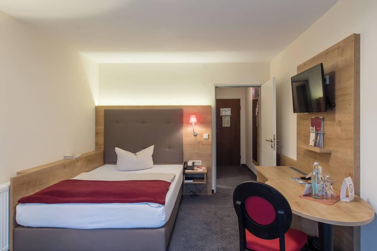 A hotel room with a bed, bedside table, lamp, TV, and a desk with a chair.