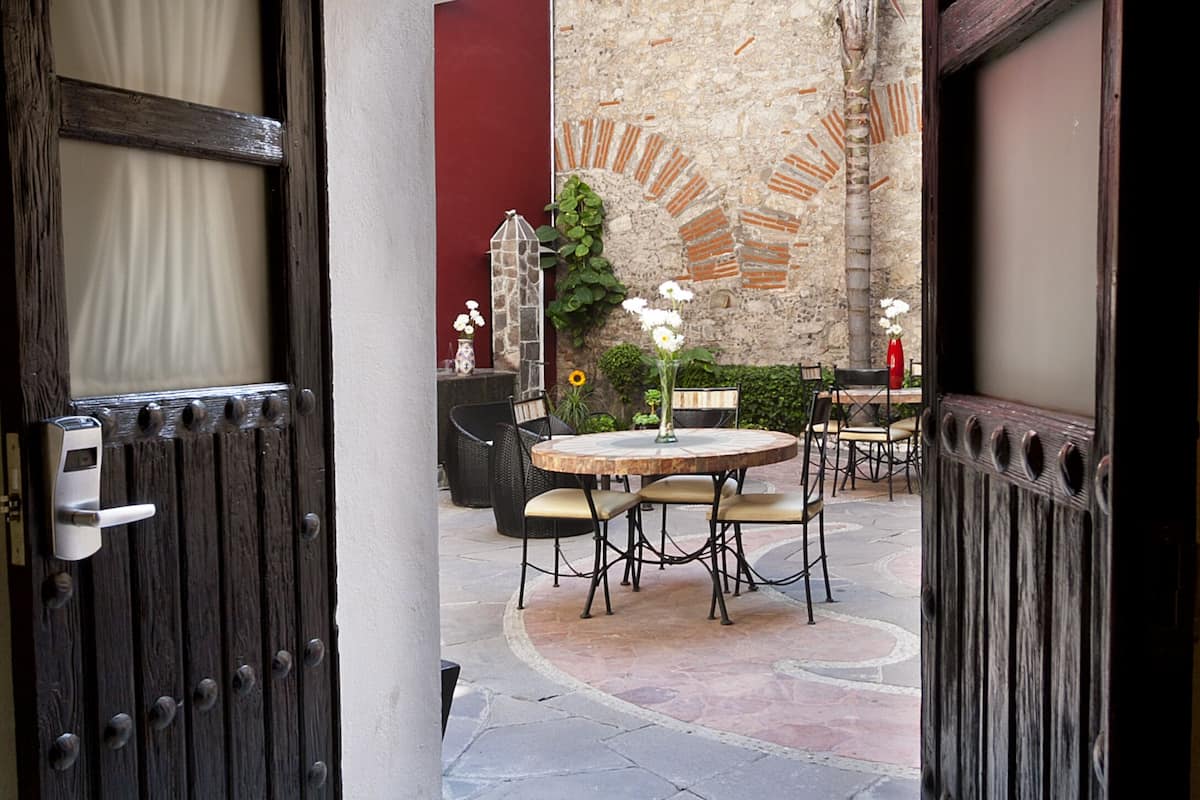 Puerta abierta que da a un patio con una mesa redonda y sillas, un pequeño jardín y un muro rojo al fondo.