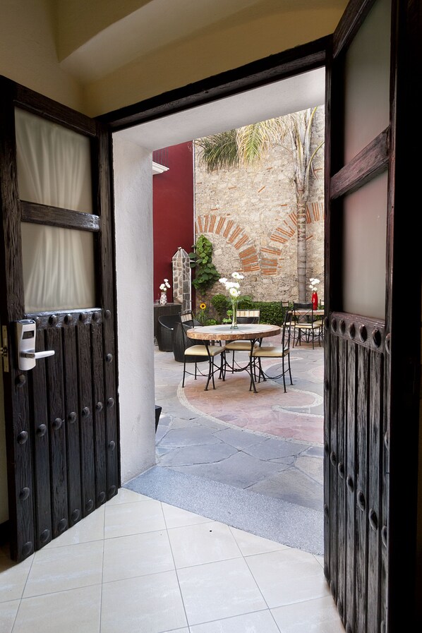 Puerta abierta que da a un patio con una mesa redonda y sillas, un pequeño jardín y un muro rojo al fondo.