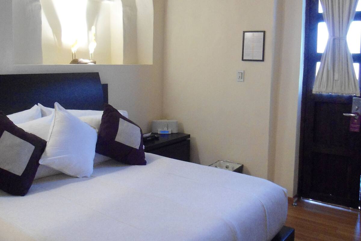 Una cama bien hecha con almohadas blancas y oscuras, una mesita de noche con lámpara y una puerta con cortinas.
