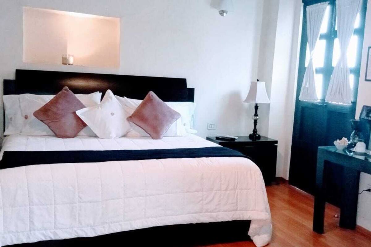 Una cama bien hecha con cabecera blanca y negra, una mesita de noche con lámpara, y un escritorio con espejo y varios objetos.