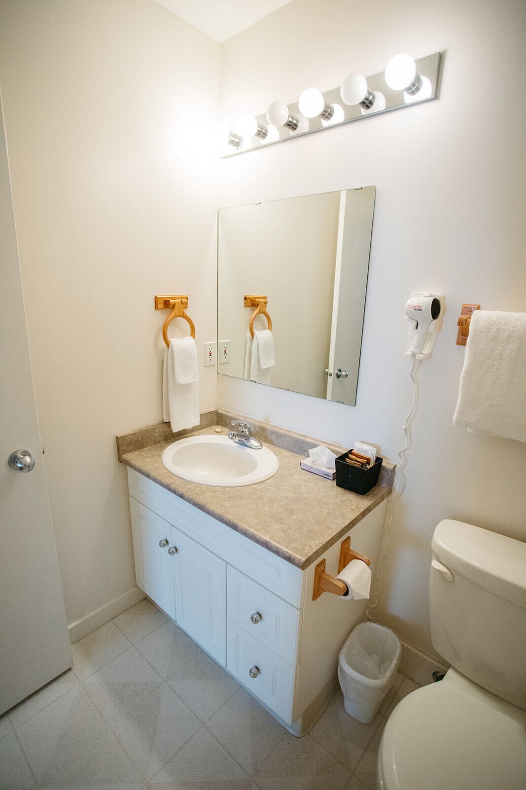 A bathroom with a white sink, mirror, and vanity, a toilet, and a trash can.