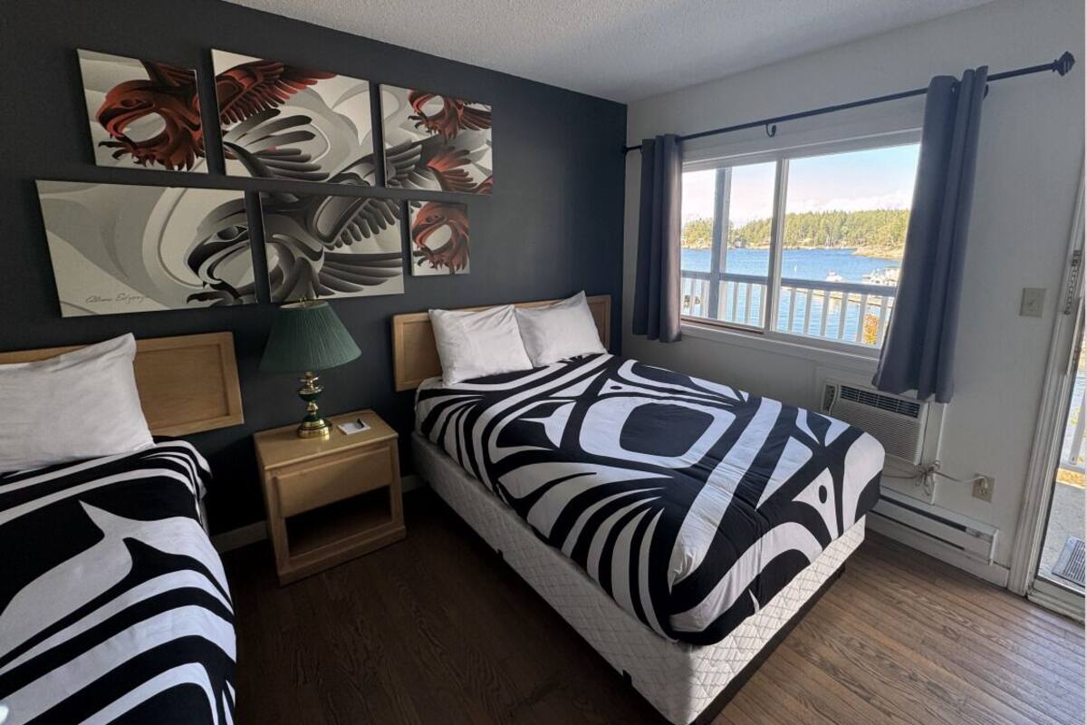Habitación de hotel con dos camas, una mesita de noche con lámpara, una obra de arte en la pared y una ventana con vista al agua.