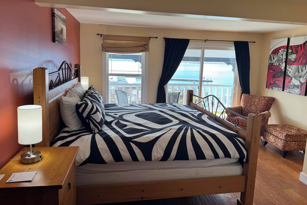 Un dormitorio con cama, mesita de noche, silla y balcón con vistas al mar.