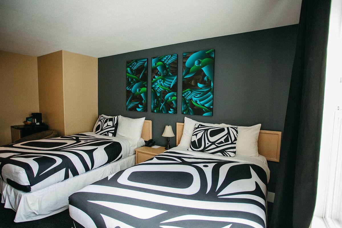 Habitación de hotel con dos camas, cada una con colcha de diseño blanco y negro. Dos cuadros adornan las paredes sobre las camas, y una pequeña mesita de noche con una lámpara y un teléfono.