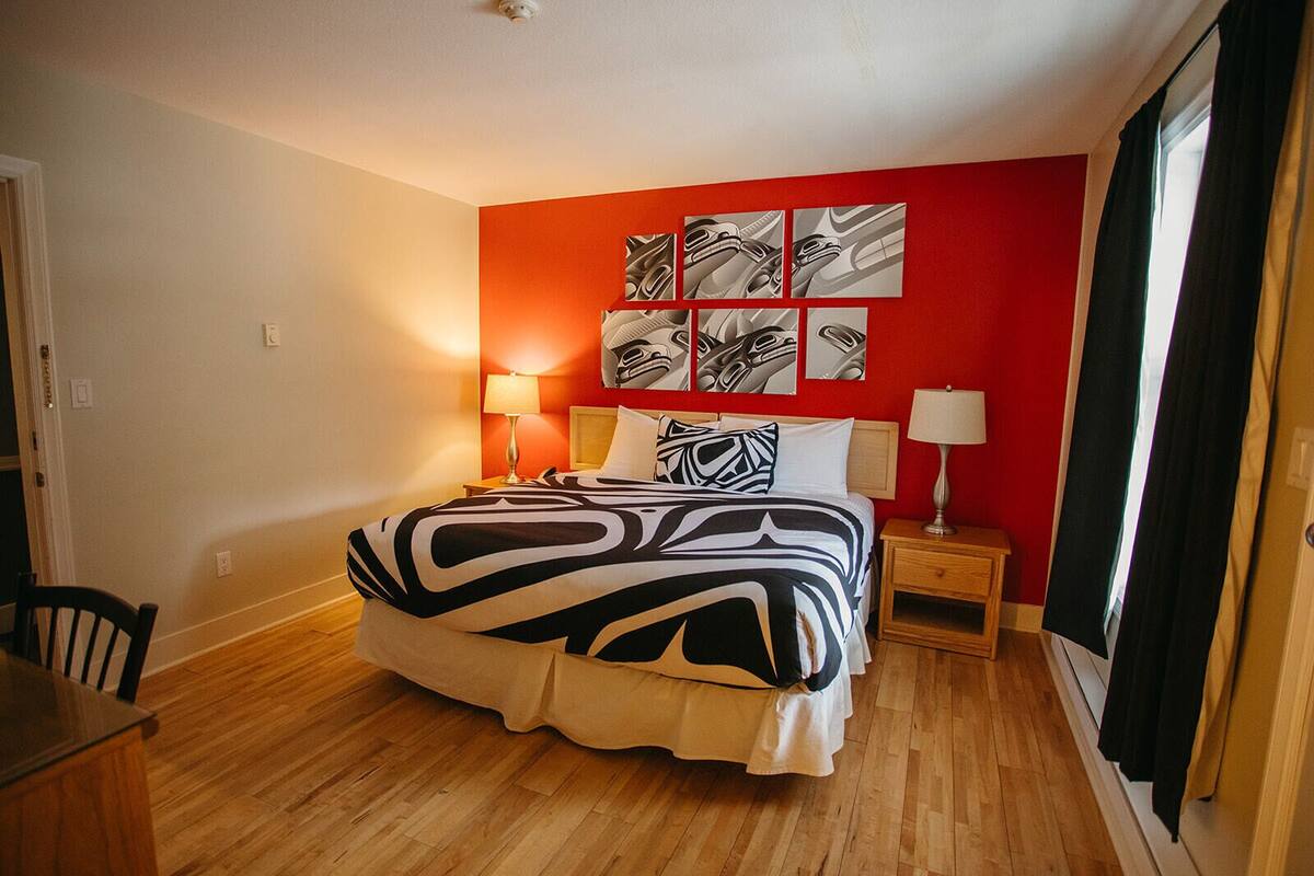 Habitación de hotel con una cama grande, dos mesitas de noche con lámparas, una pared de acento roja y un ventanal con cortinas.