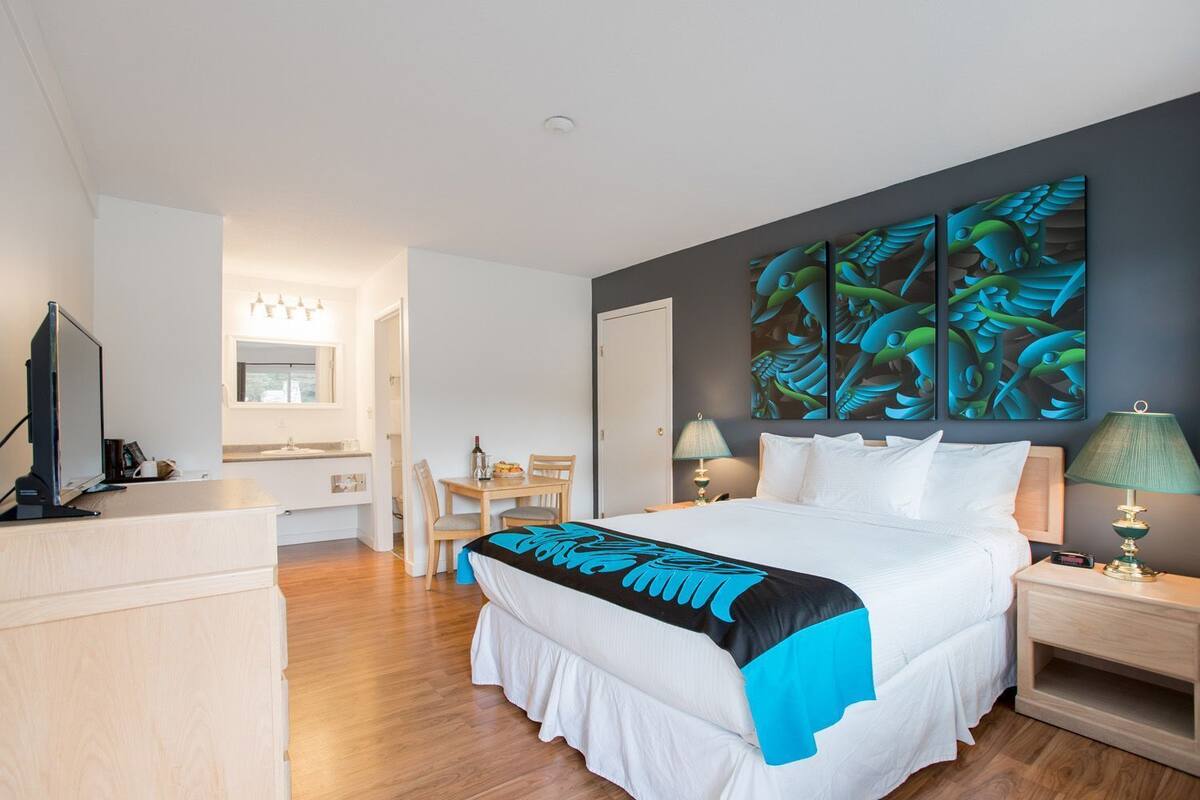 Una habitación de hotel con cama, escritorio, silla, televisor y obras de arte en la pared.