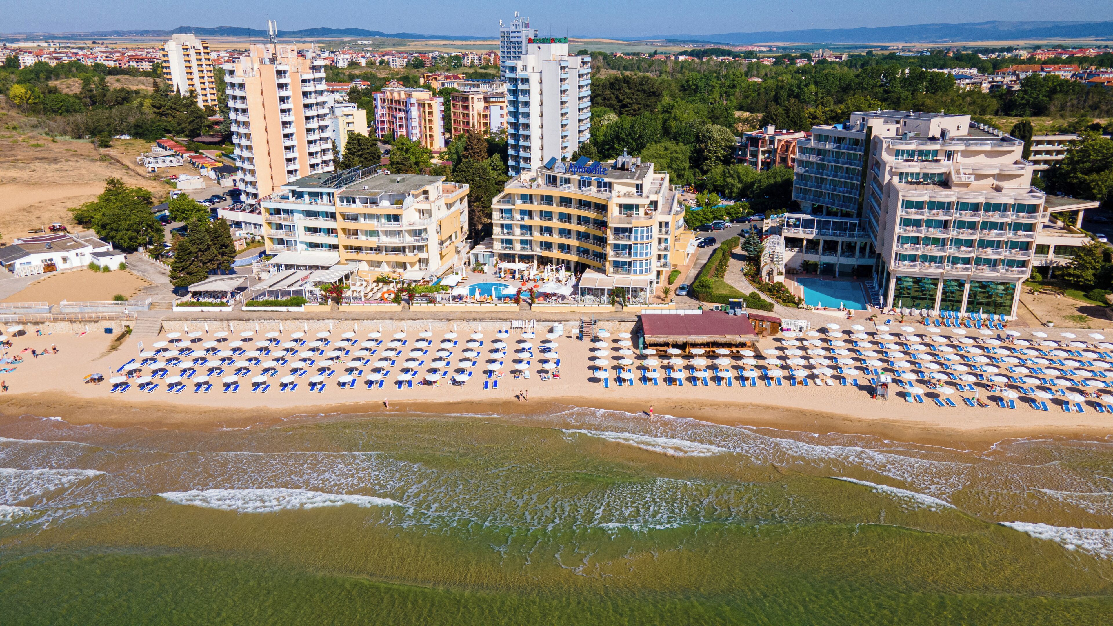 Un hotel en la costa con varias edificaciones, una playa con tumbonas y una piscina.