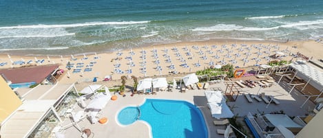 A pool area with lounge chairs and umbrellas, a beach with umbrellas and lounge chairs, and the ocean in the background.