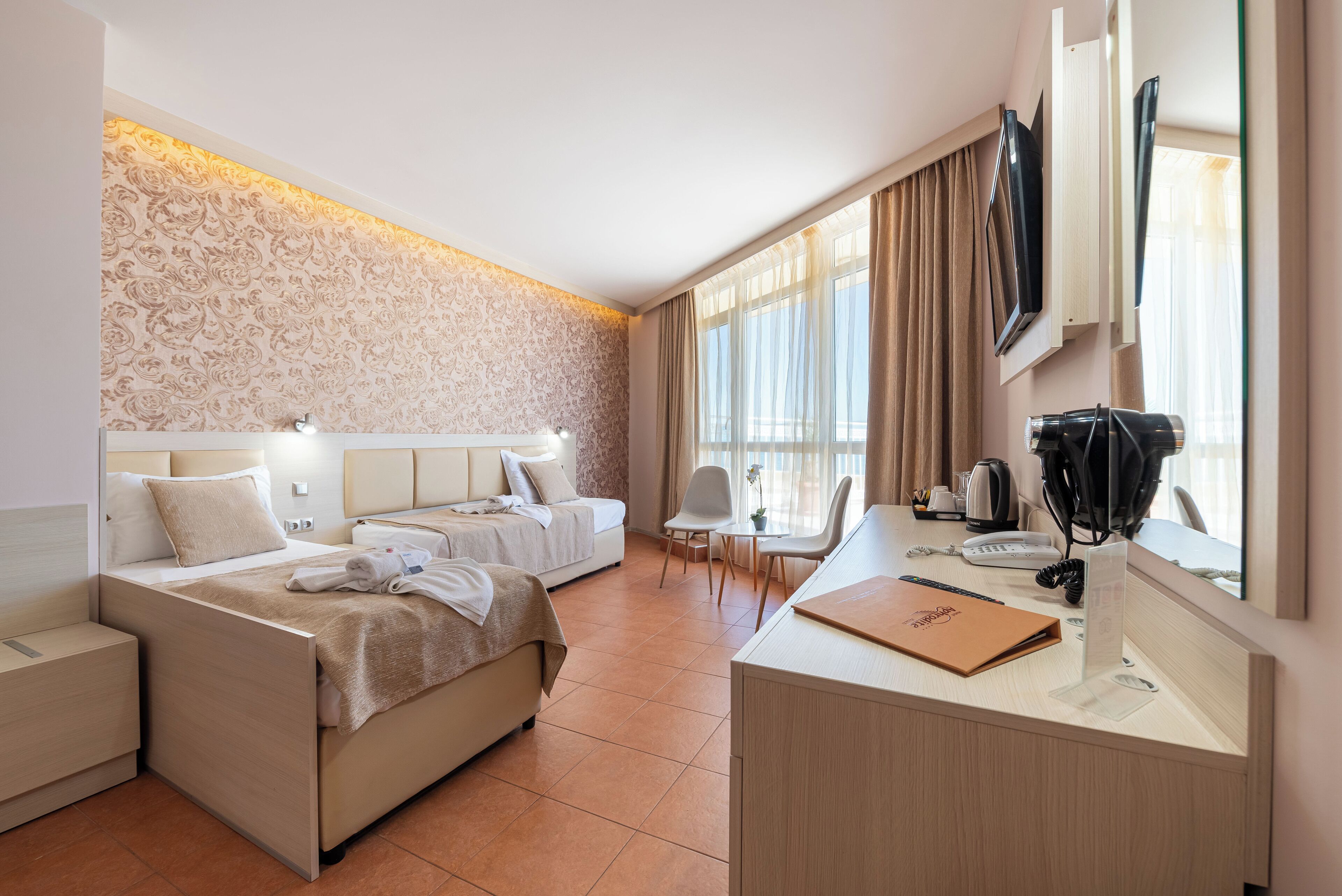 A hotel room with two beds, a desk with a telephone and a laptop, a TV mounted on the wall, and a window with curtains.