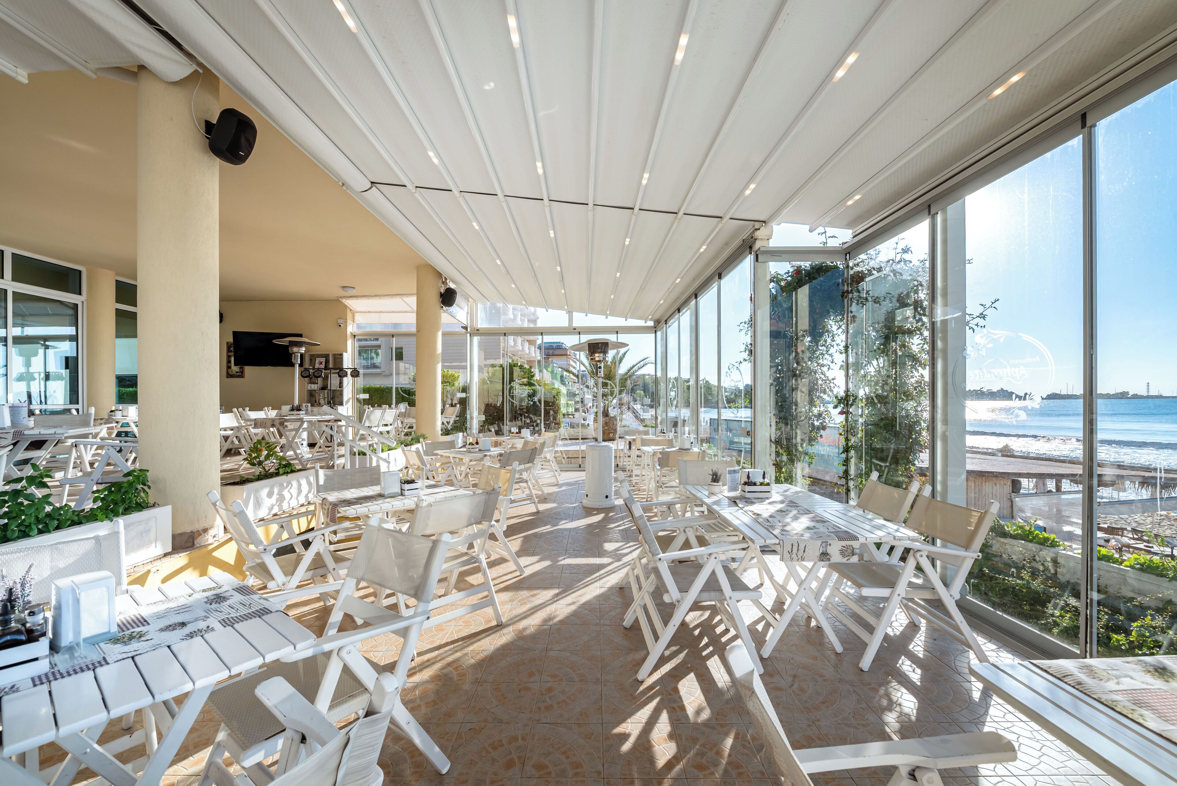 Un espacio comedor al aire libre con mobiliario blanco y ventanas amplias con vistas al mar.