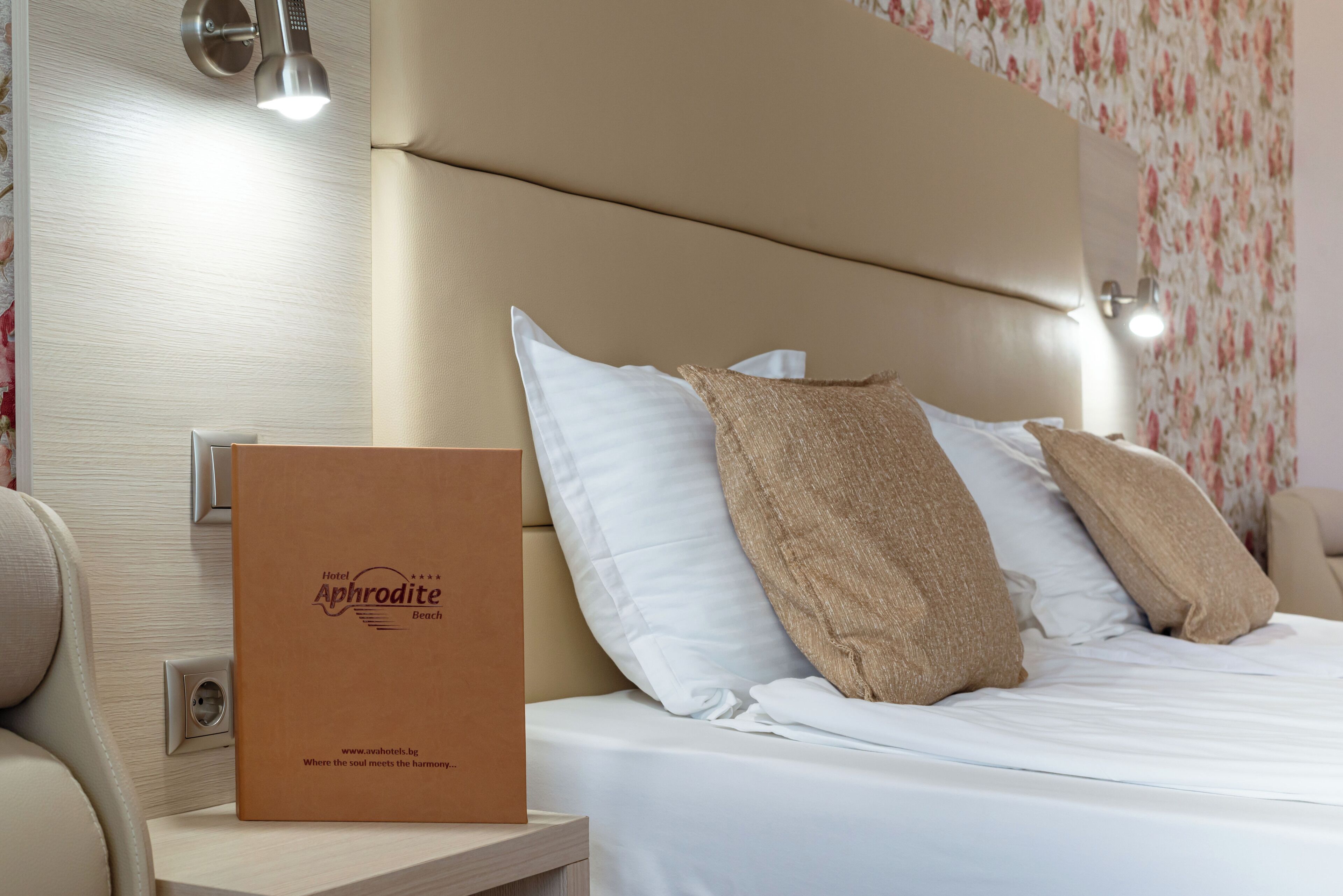 A neatly made bed with white and beige pillows, a brown hotel brochure on the nightstand, and a wall-mounted light fixture.