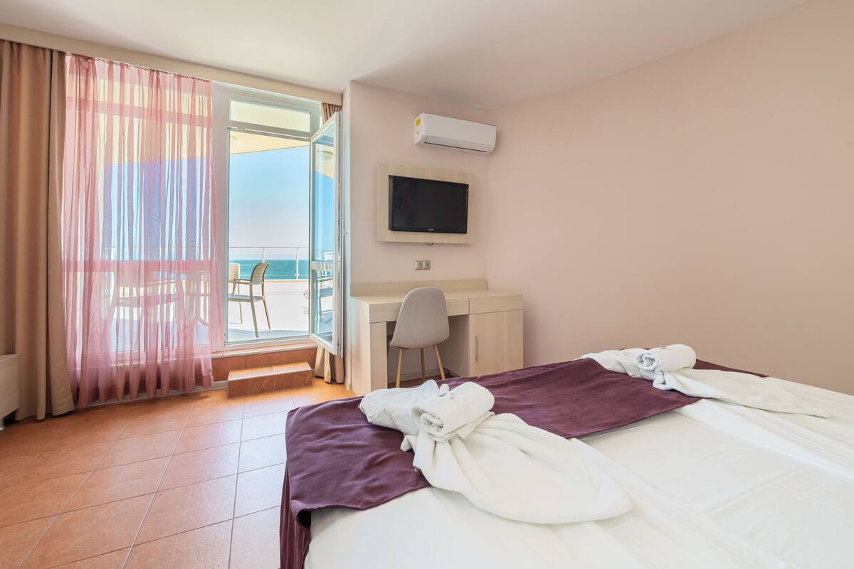 A hotel room with a bed, a desk, a chair, a TV, and a view of the sea.