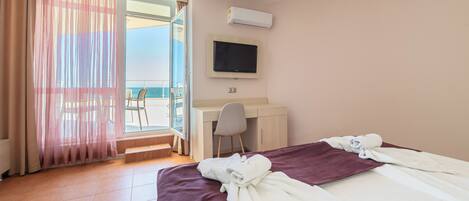 A hotel room with a bed, a desk, a chair, a TV, and a view of the sea.