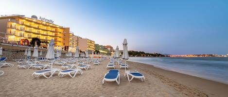 A beach with sun loungers and umbrellas, a hotel named 'Aphrodite' in the background, and a calm sea.