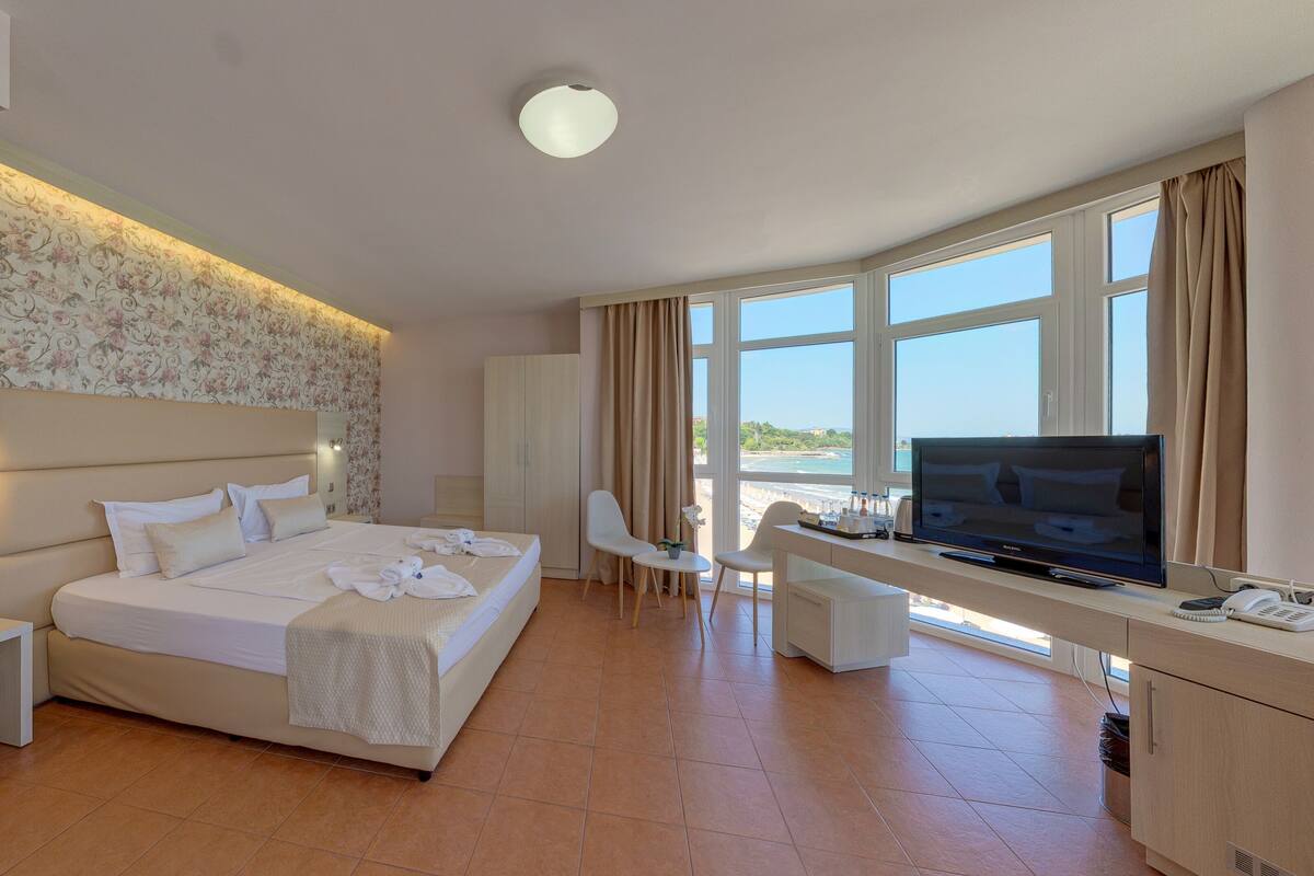 A hotel room with a large bed, a desk with a TV, a chair, and a view of the sea.