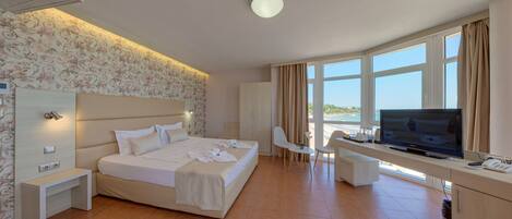A hotel room with a large bed, a desk with a TV, a chair, and a view of the sea.