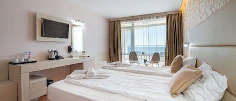 A hotel room with two beds, a desk, a TV, and a view of the sea.